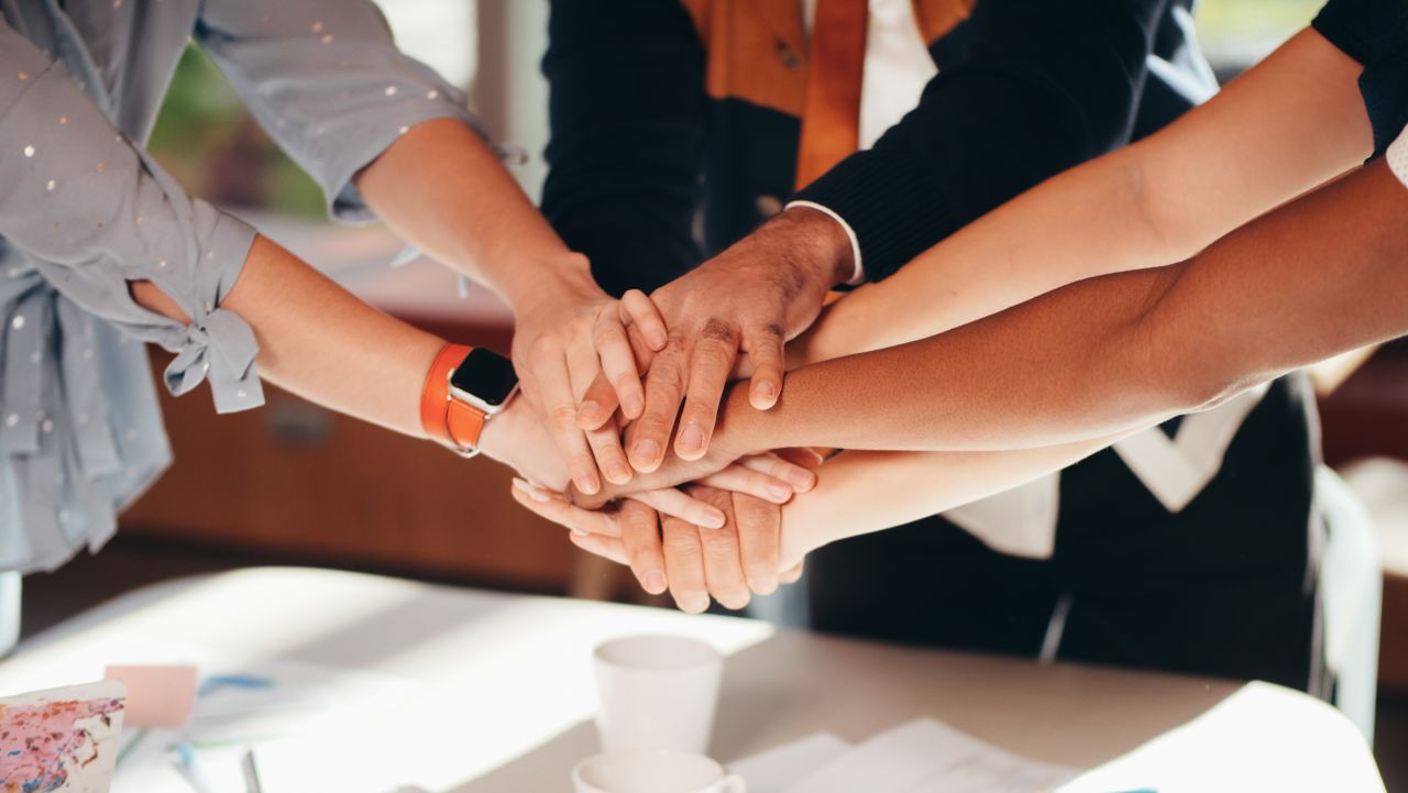 gallery-1 A close-up shot of diverse team members stacking hands over a white table, symbolizing unity and collaboration.